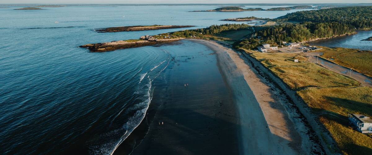 An aerial view of a coastal area with a sandy beach, a road, and a small settlement with buildings.