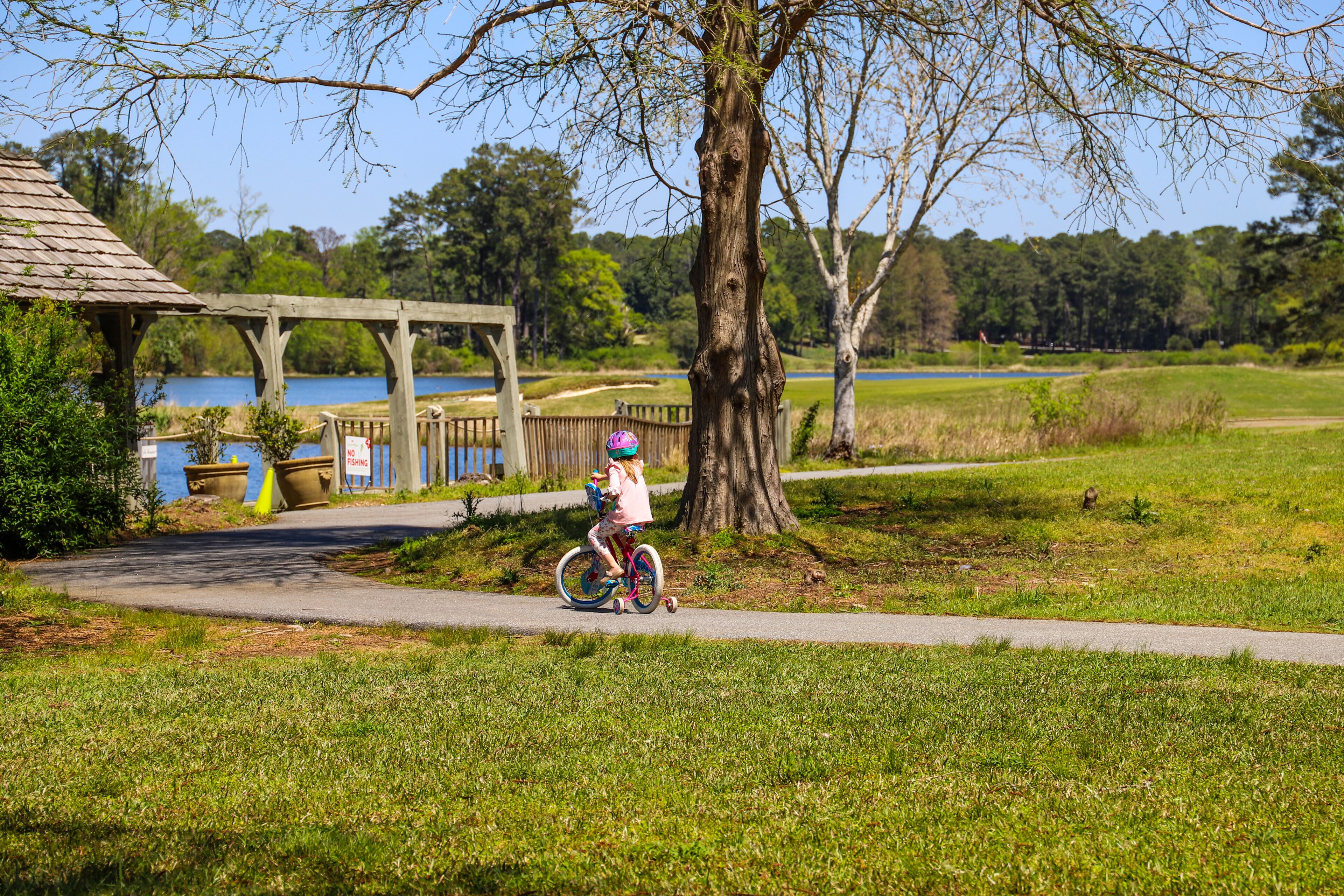a little girl riding a bike on a smooth winding footpath in the garden surrounded by lush green trees, grass and plants near a blue lake with blue sky at Callaway Gardens in Pine Mountain Georgia USA