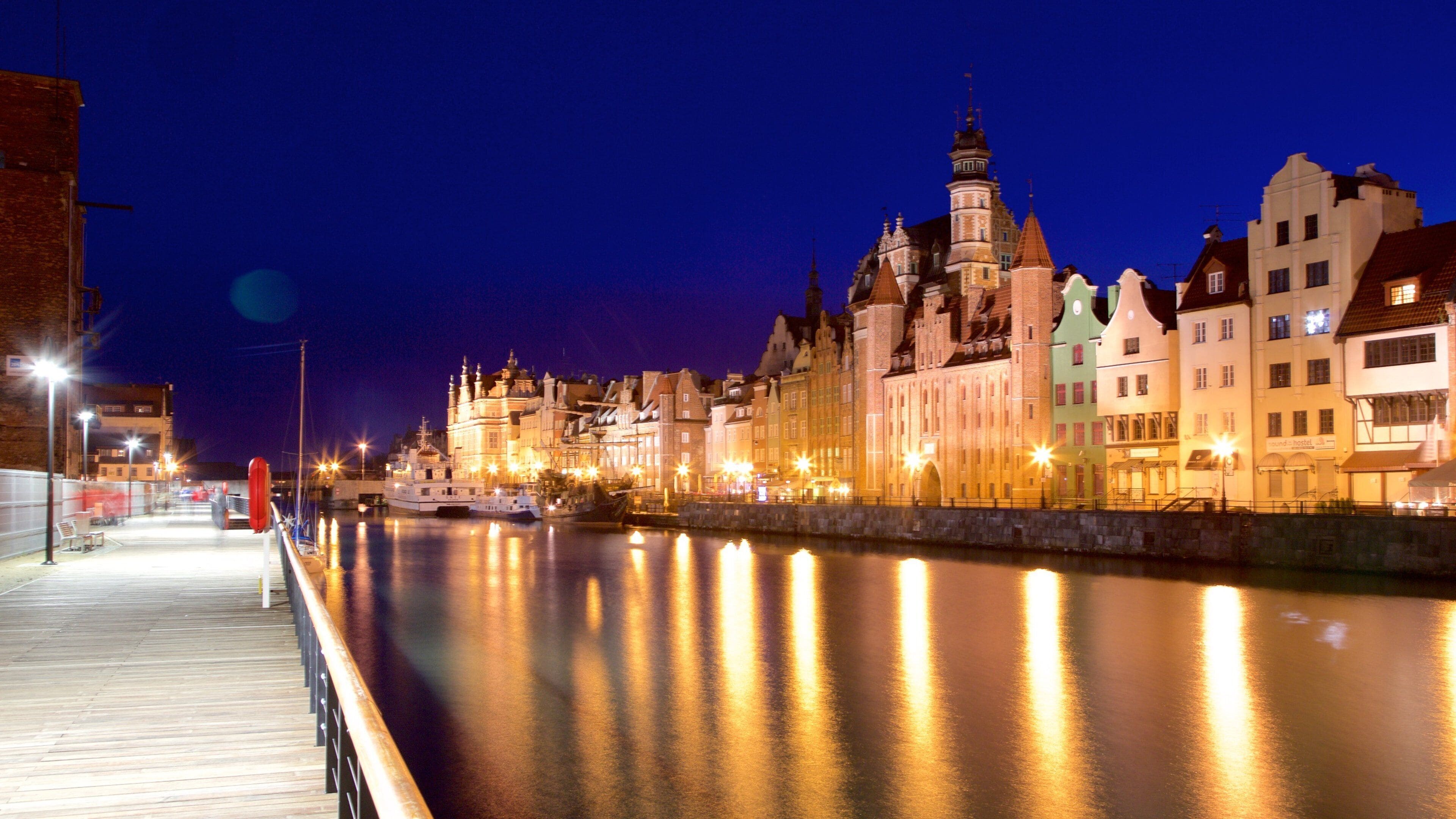A nighttime view of a river with a wooden walkway and illuminated buildings along the riverbank.