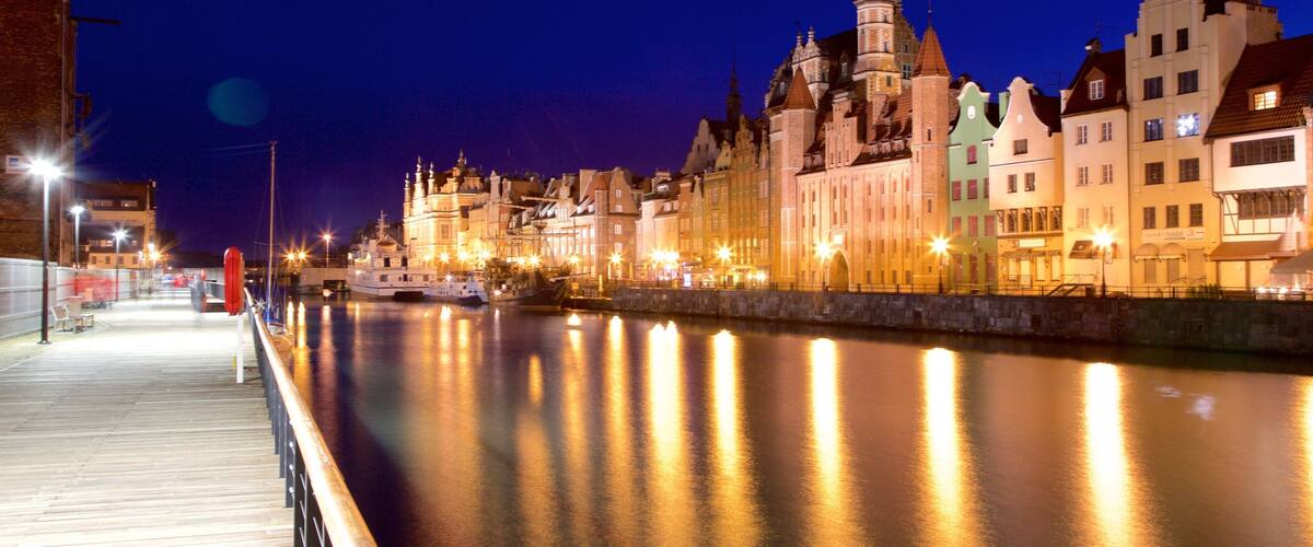 A nighttime view of a river with a wooden walkway and illuminated buildings along the riverbank.
