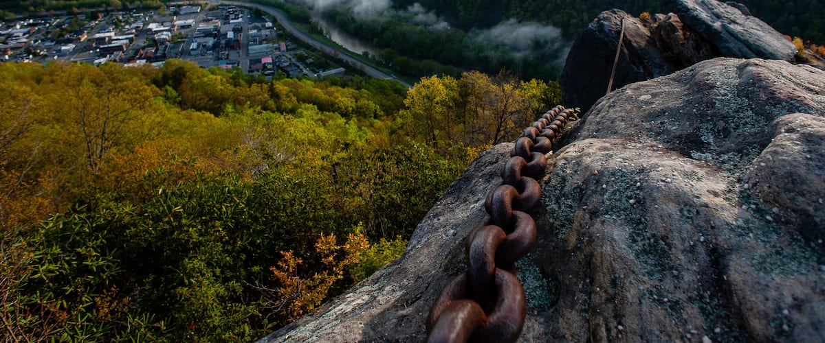A mountainous landscape with a town in the valley, a winding road, and a chain of rusty metal rings on a rock.
