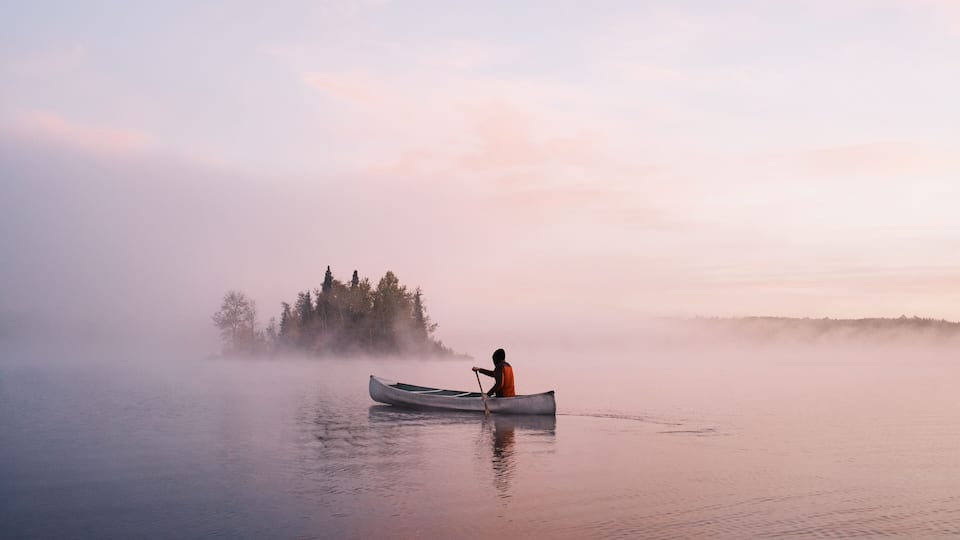 A calm lake with a misty forest in the background and a person in a canoe.