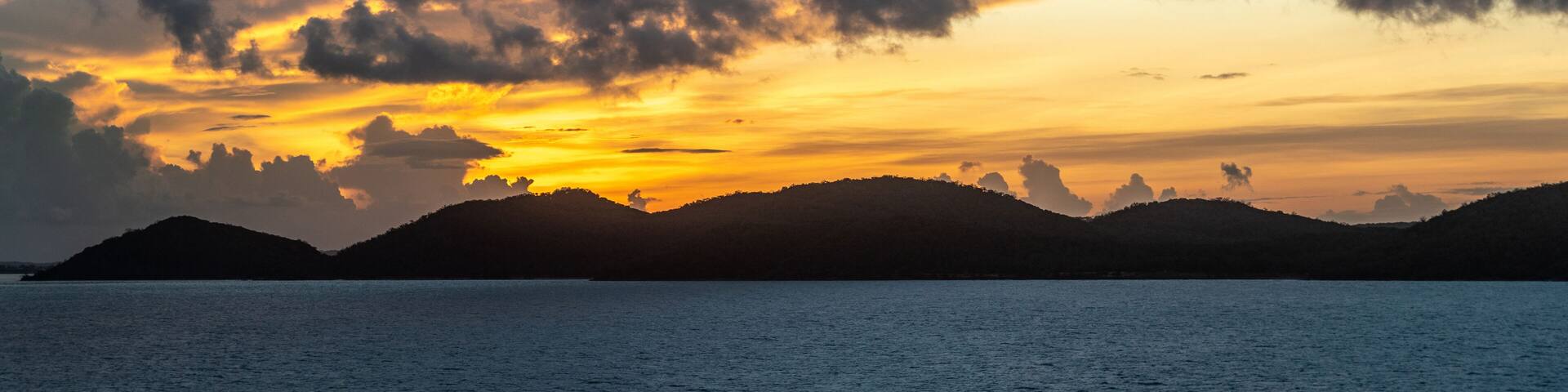 Thursday Island, Australia - February 20, 2019: Pre-sunrise shot over Torres Strait Islands Archipelago shows dark blue clouds in yellow and red light hanging over black island hills in very dark blue