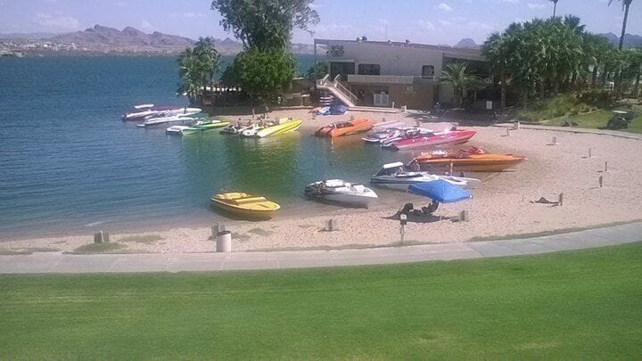 Un puerto deportivo con diversas embarcaciones de colores amarradas a una playa arenosa, rodeado de vegetación y un edificio al fondo.