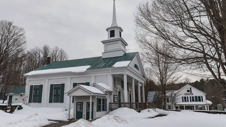 A white church with a green roof and a steeple, surrounded by snow and leafless trees.