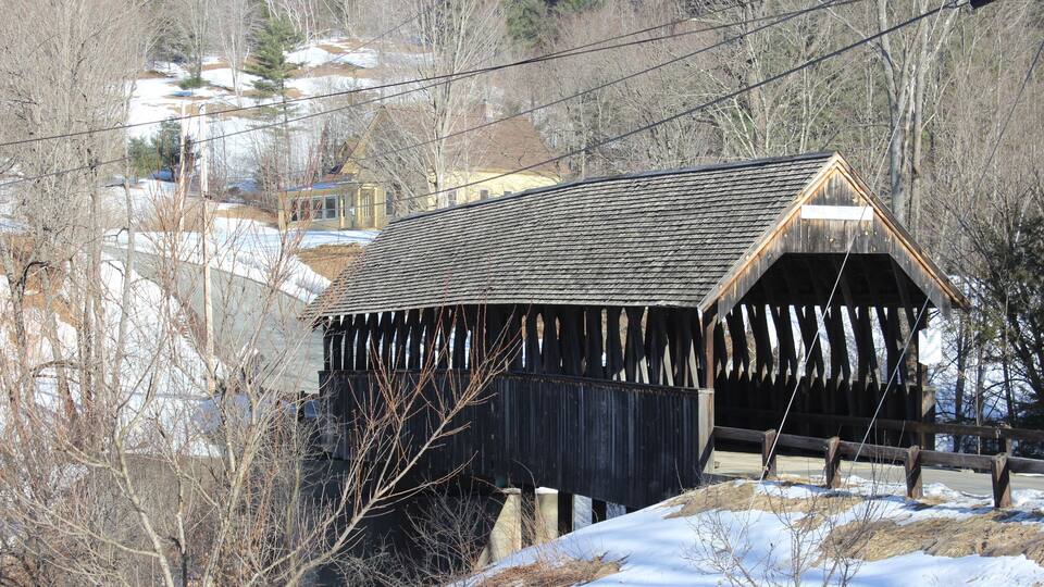 A wooden covered bridge with a steep roof, surrounded by snow-covered ground and leafless trees.