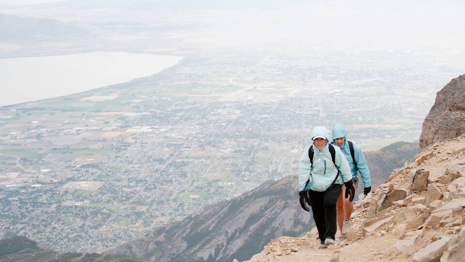 Two hikers on a rocky mountain path with a city and lake in the background.