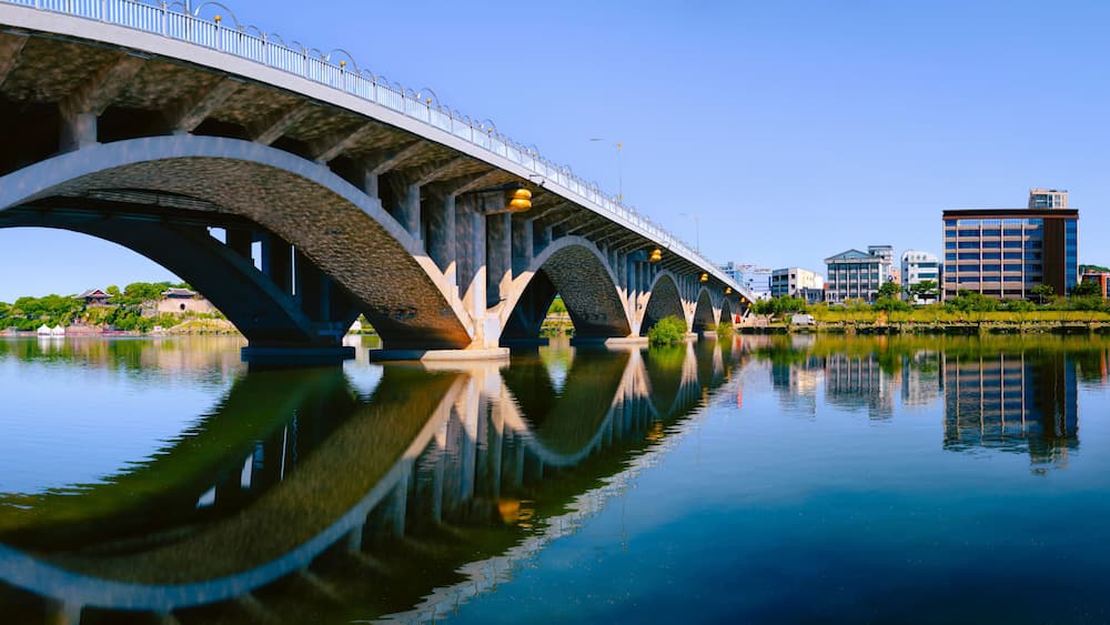 A bridge over a calm river with clear reflections, surrounded by urban buildings.