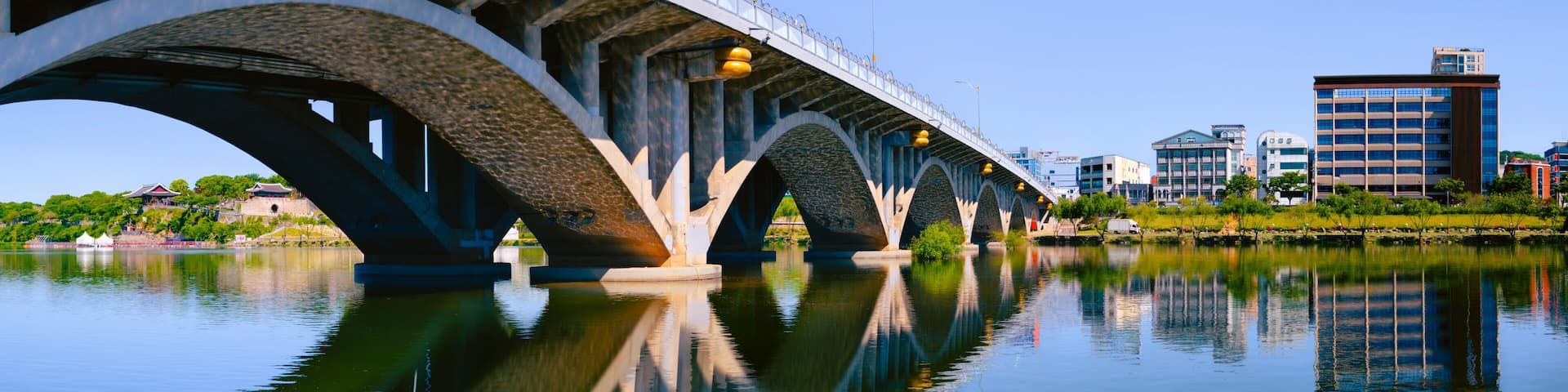 A bridge over a calm river with clear reflections, surrounded by urban buildings.