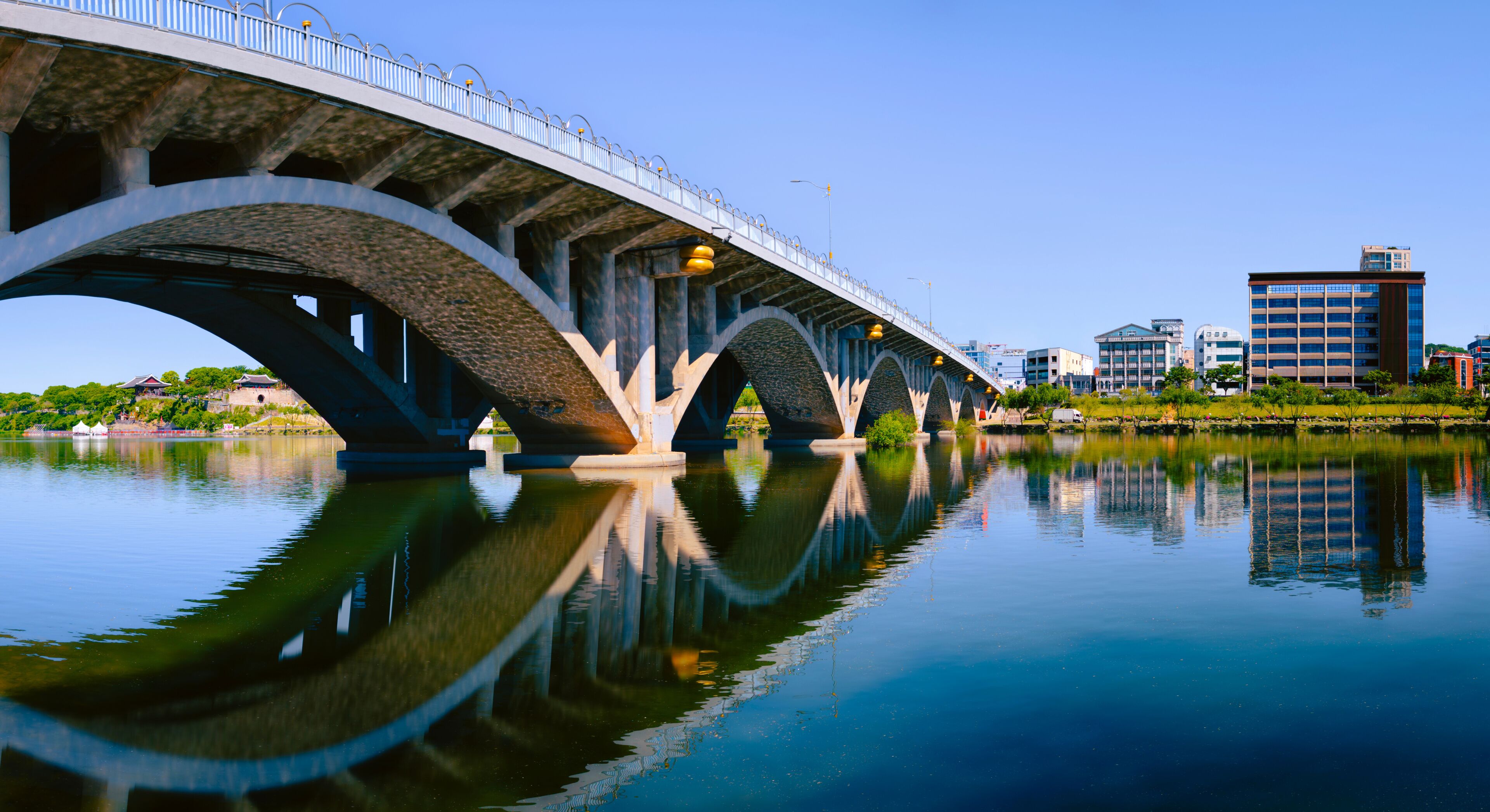 A bridge over a calm river with clear reflections, surrounded by urban buildings.