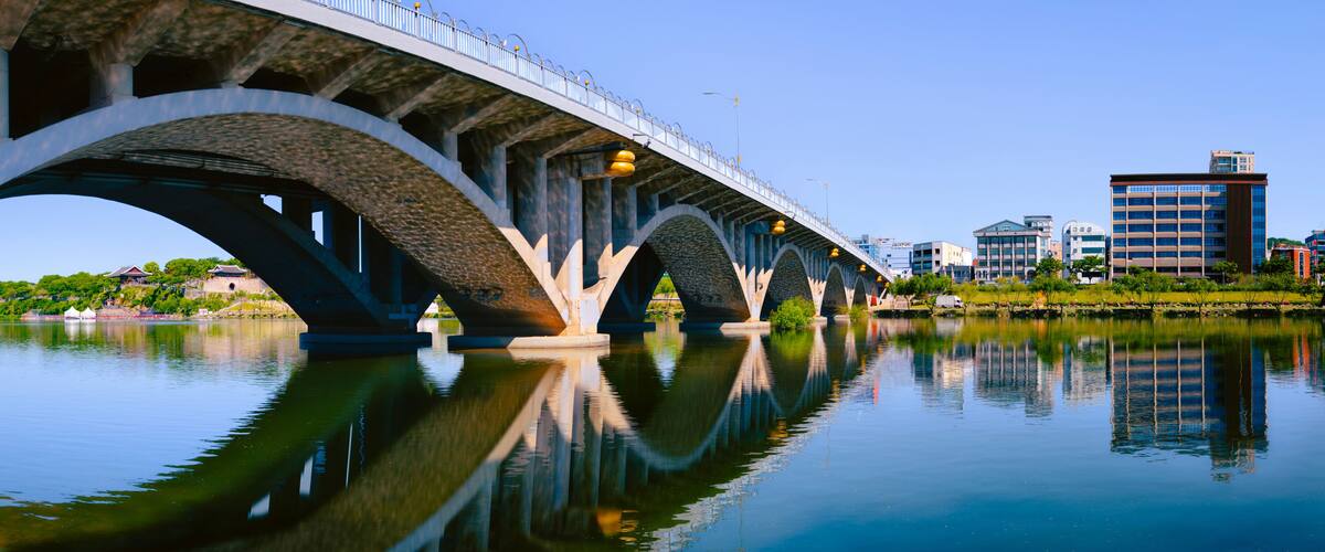 A bridge over a calm river with clear reflections, surrounded by urban buildings.