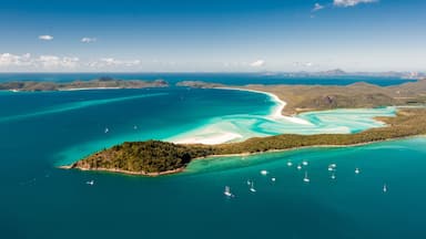 Vue aérienne d’une zone côtière aux eaux turquoise et limpides, avec une petite île couverte d’une végétation luxuriante et des montagnes lointaines sous un ciel dégagé.