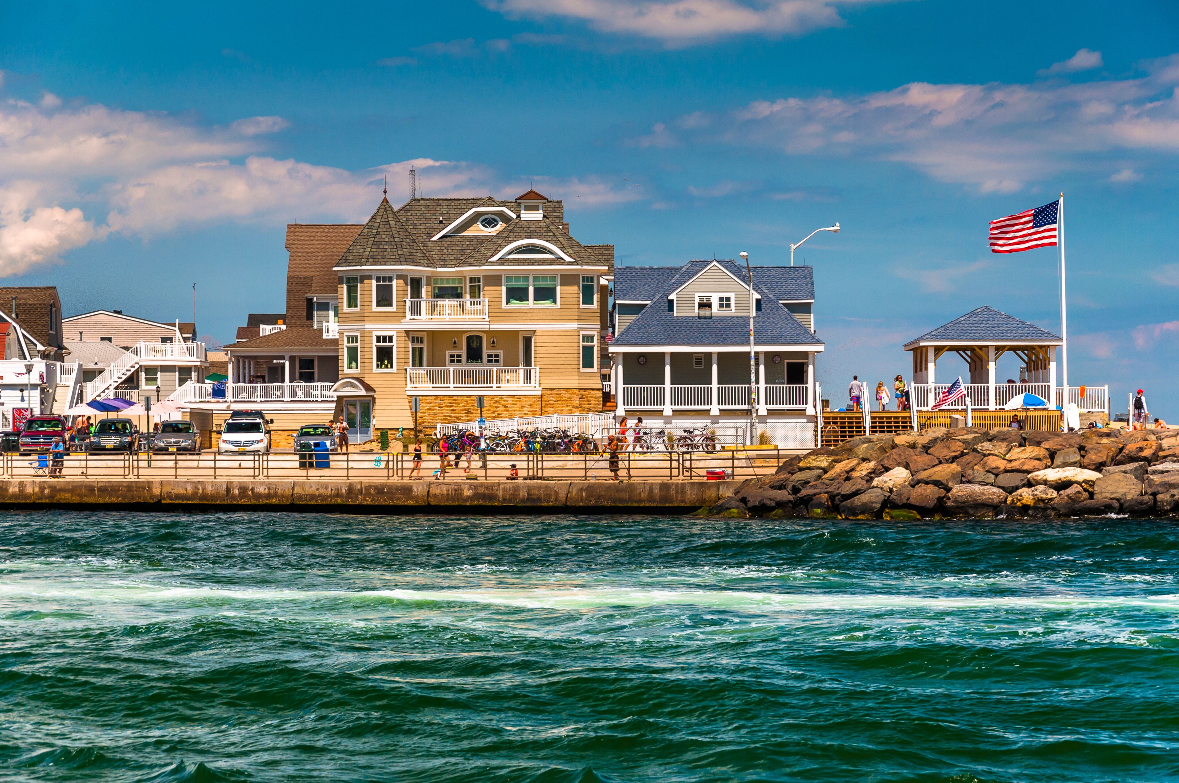 A coastal town with a prominent yellow house, a gazebo with an American flag, and a rocky breakwater.