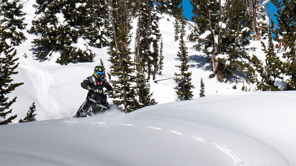 A snowmobile rider navigating a snow-covered terrain with trees in the background.