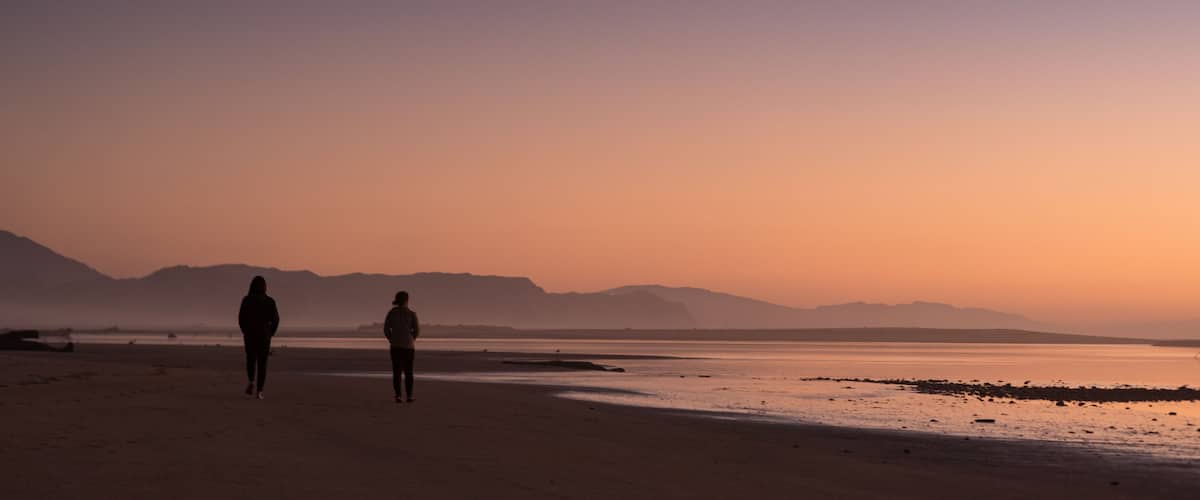 A beach with two people walking, mountains in the background, and a sunset sky.