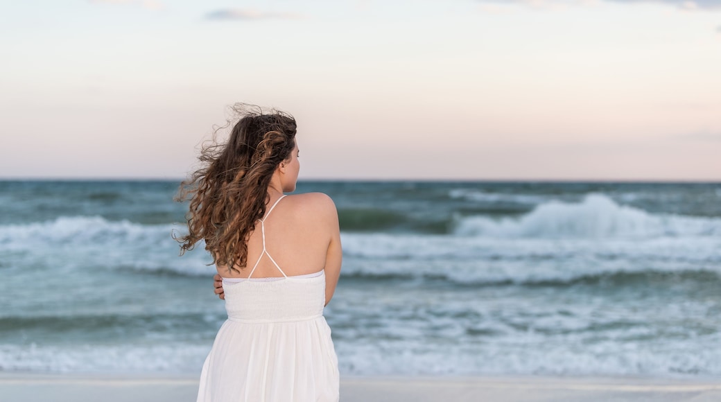 Young woman back standing in white dress on beach evening in Florida panhandle shivering arms crossed in wind by ocean waves