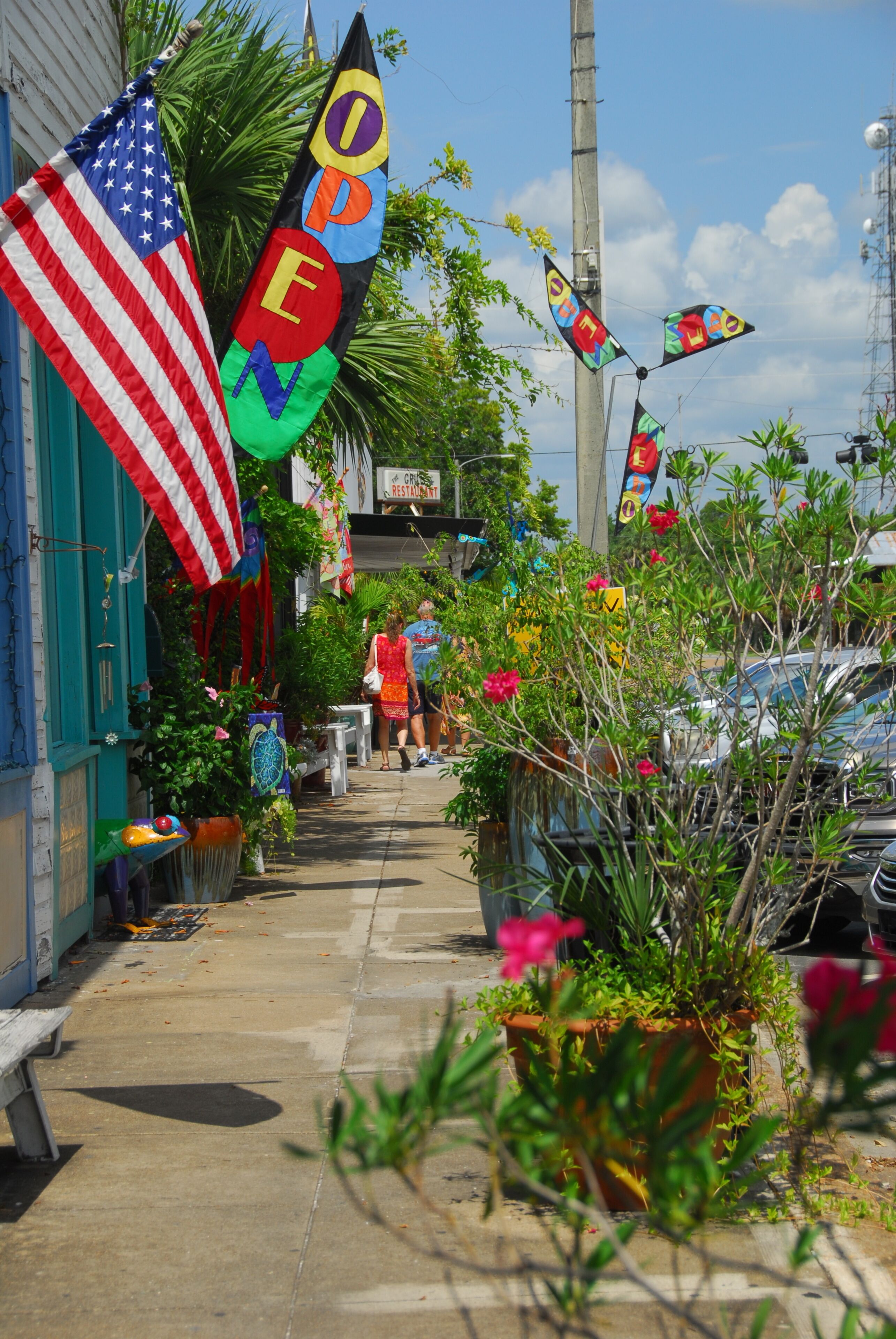 A sidewalk lined with potted plants, American flags, and colorful flags with the word 'OPEN' on them.