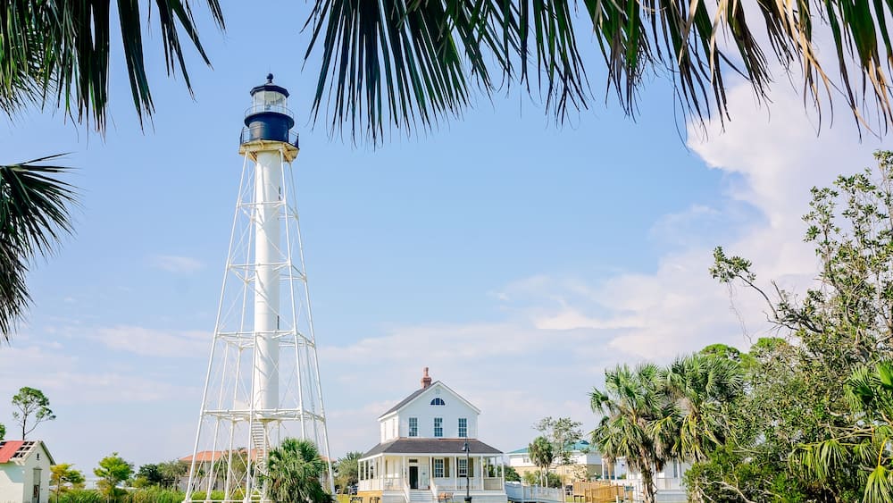 Un phare doté d’une tour blanche et d’une partie supérieure bleue, une maison blanche avec un porche, et une végétation luxuriante environnante, notamment des palmiers.