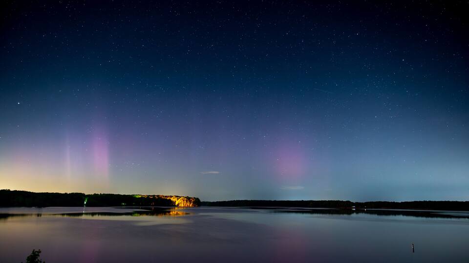 A night sky with stars and the aurora borealis over a calm lake with a distant shoreline.