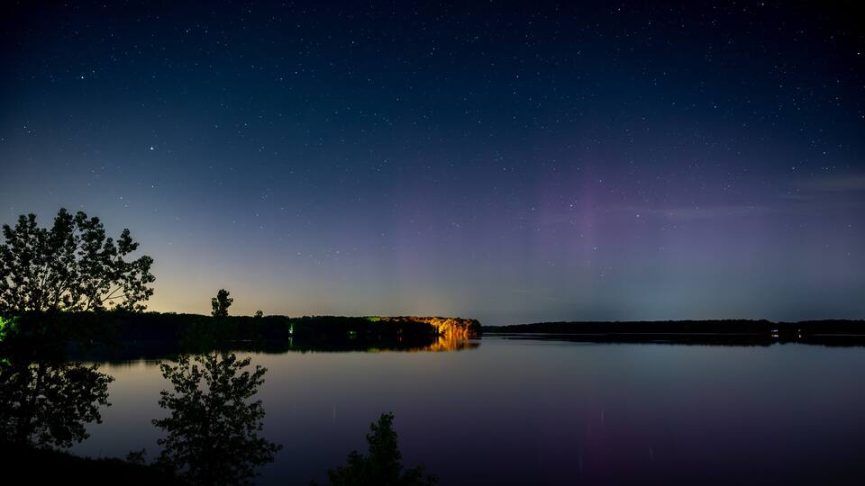 A starry night sky over a calm lake with a silhouette of a tree and a distant illuminated structure.
