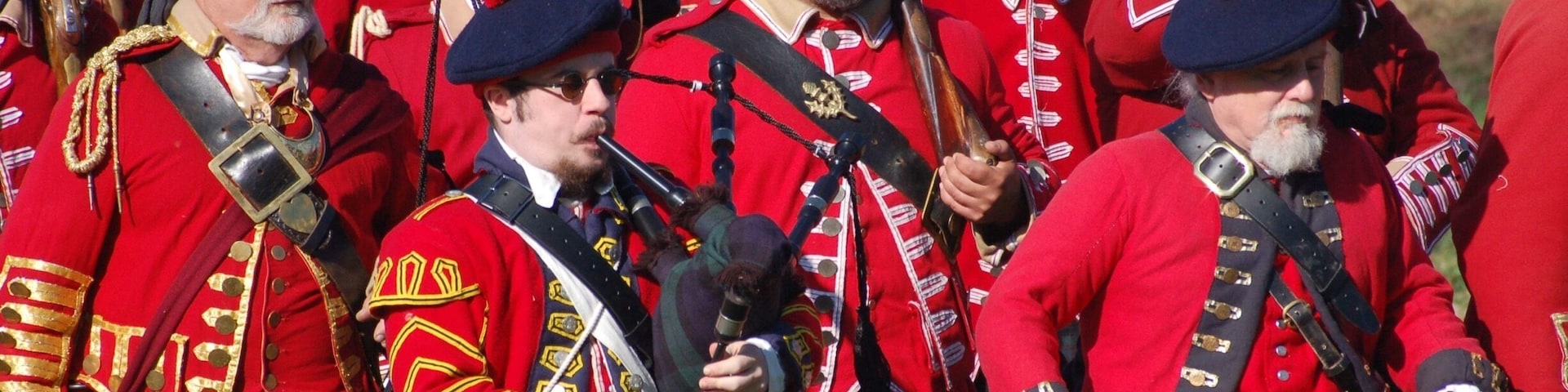 A group of men in red uniforms with gold accents, holding rifles and wearing kilts.