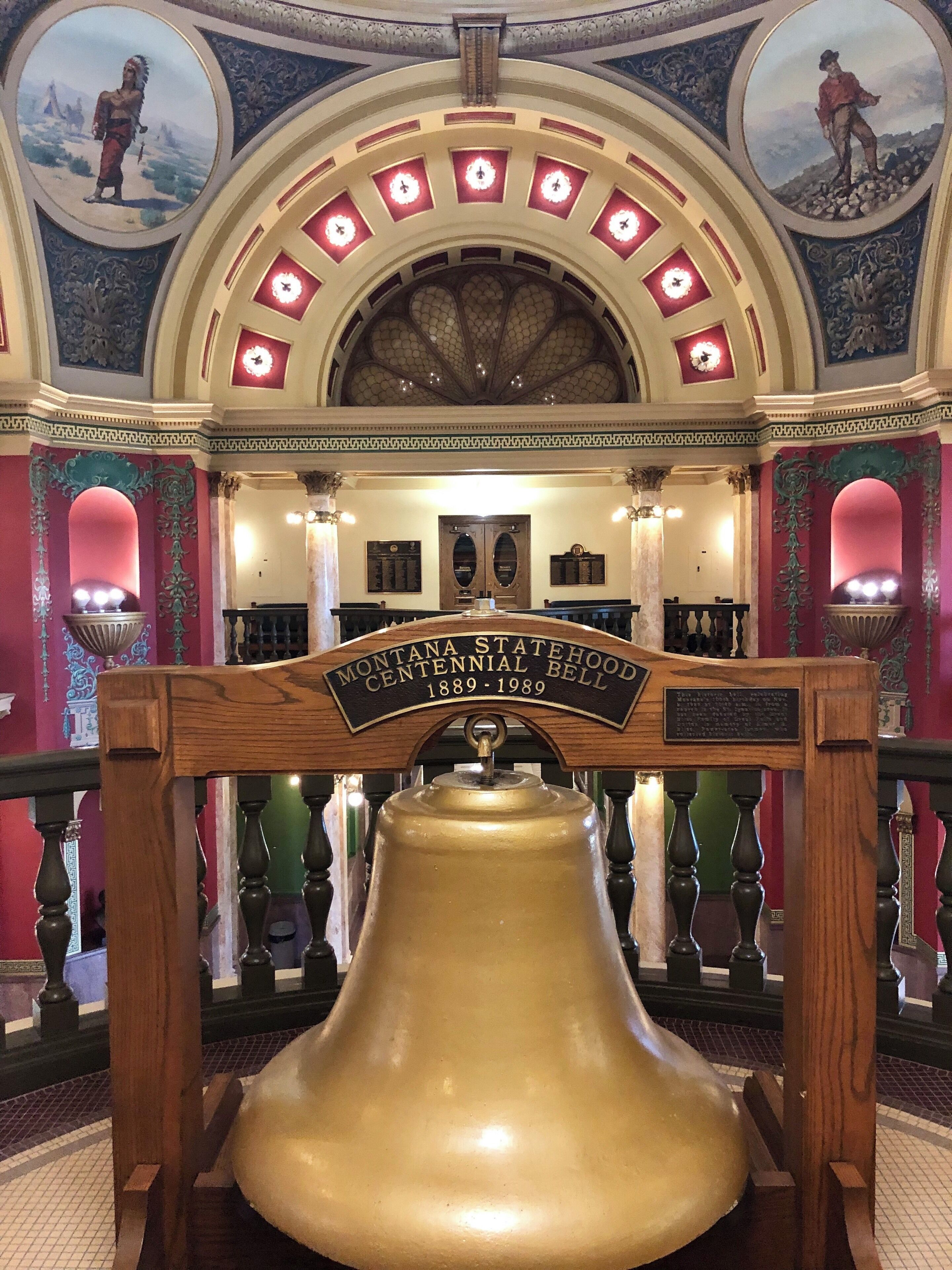 A large bell with a sign reading "Montana Statehood Centennial Bell 1889-1989" in a decorated interior.