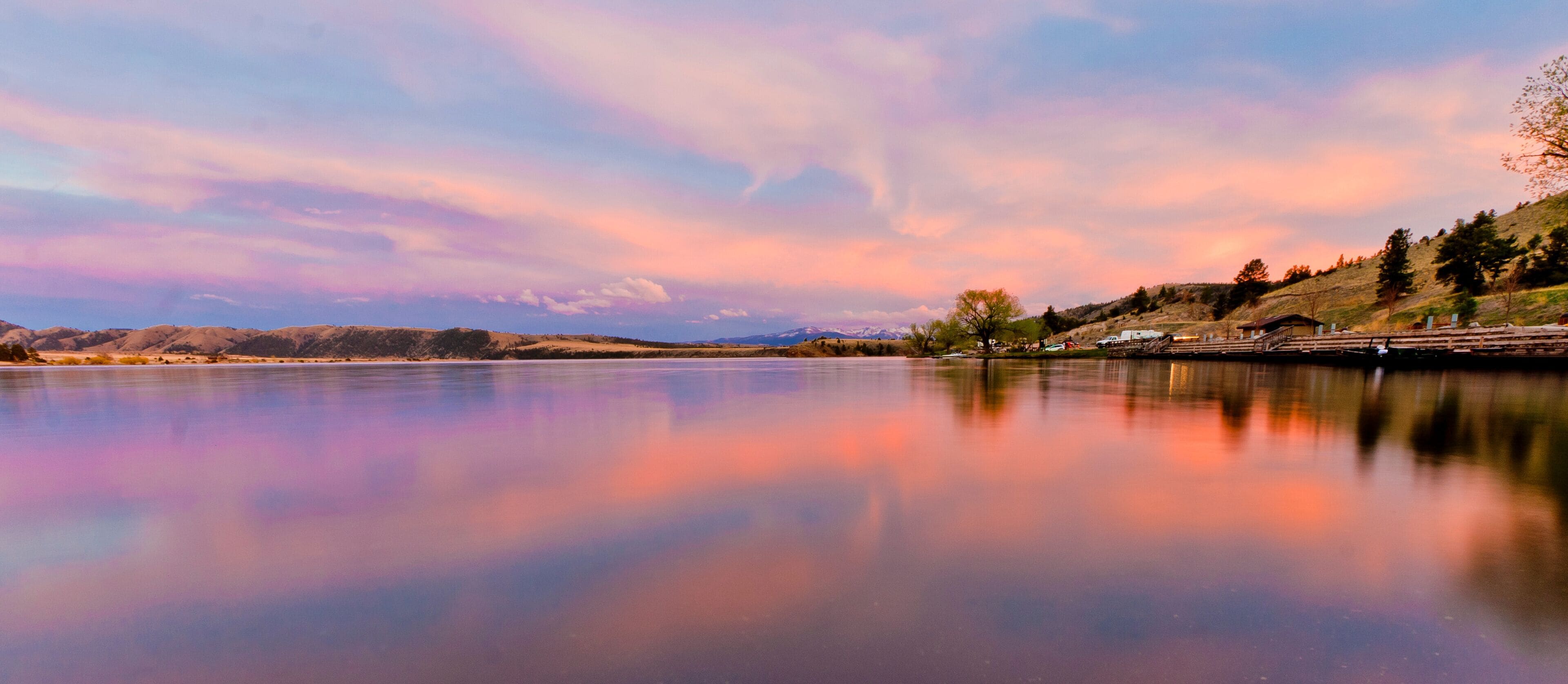 Um lago tranquilo refletindo o céu, rodeado por colinas e árvores esparsas.