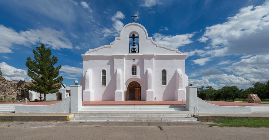 A white church with a cross on top, a bell tower, and a large tree in front.