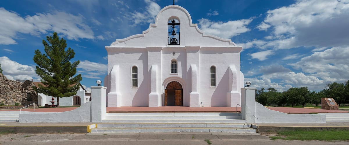 A white church with a cross on top, a bell tower, and a large tree in front.