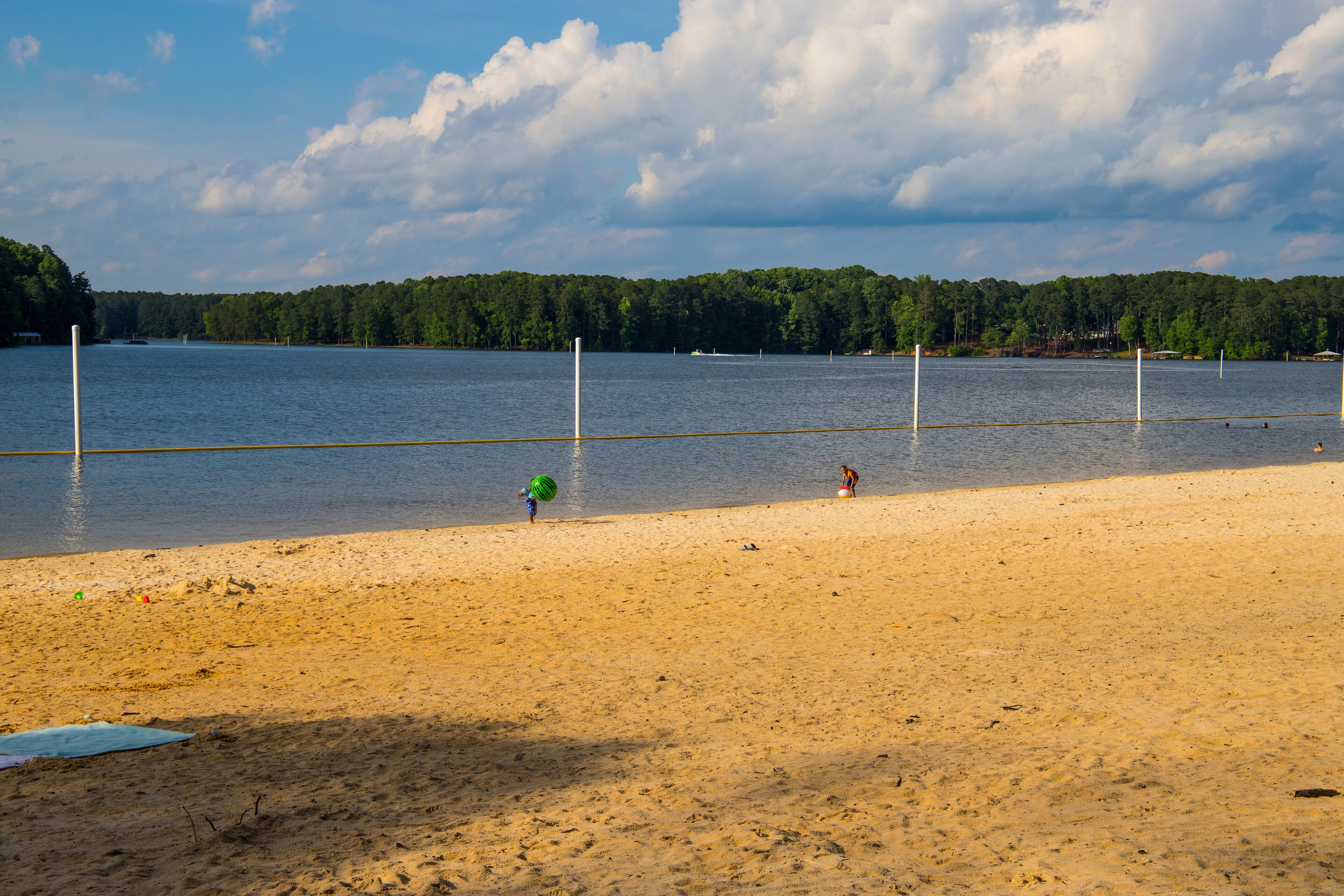 people playing in the water at the sand beach surrounded by the blue waters of Lake Acworth with lush green trees, grass and plants with blue sky and clouds at Proctor Landing Park in Acworth Georgia 