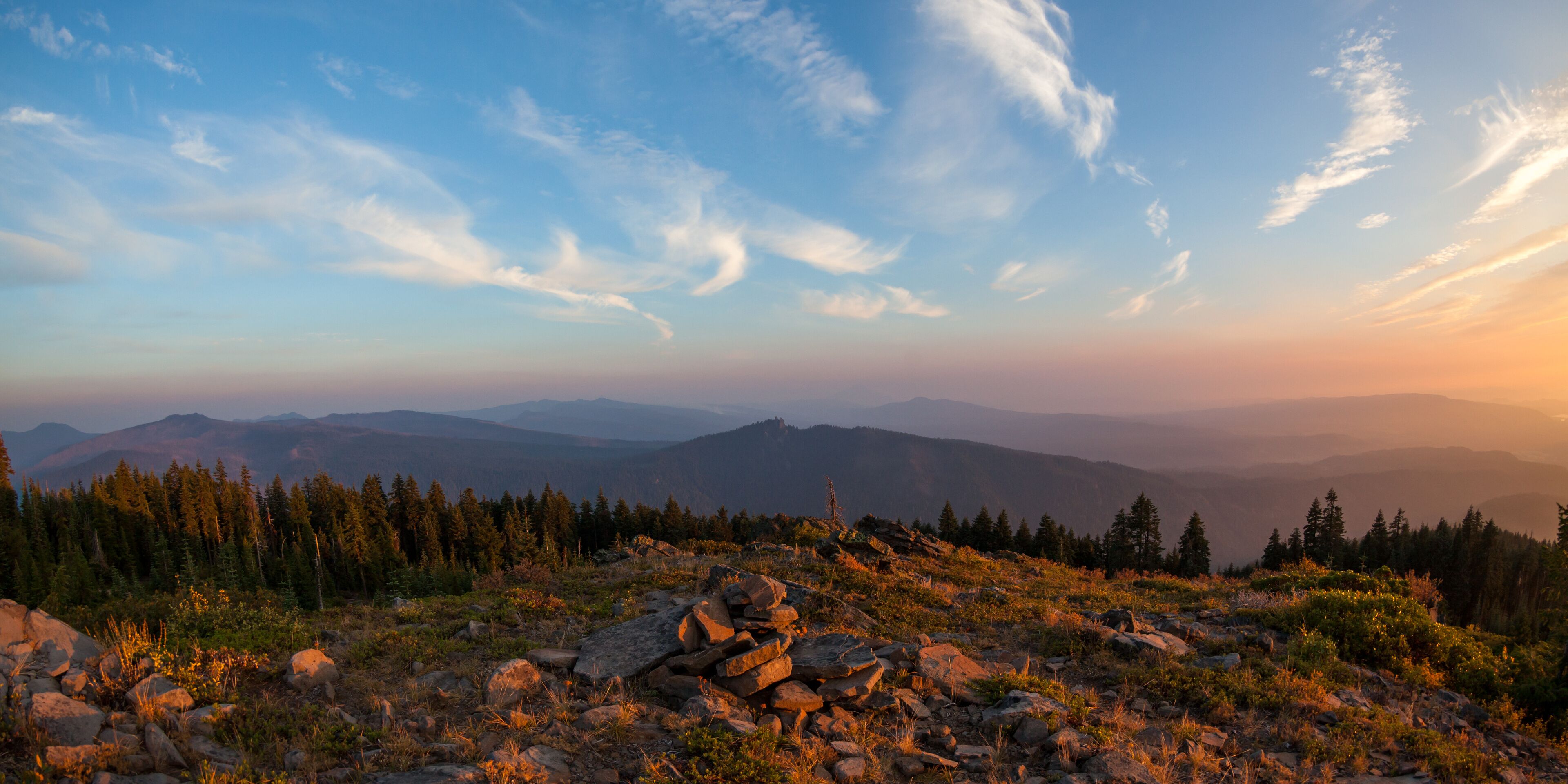 A mountainous landscape with rocky terrain, dense forests, and a clear sky with scattered clouds.