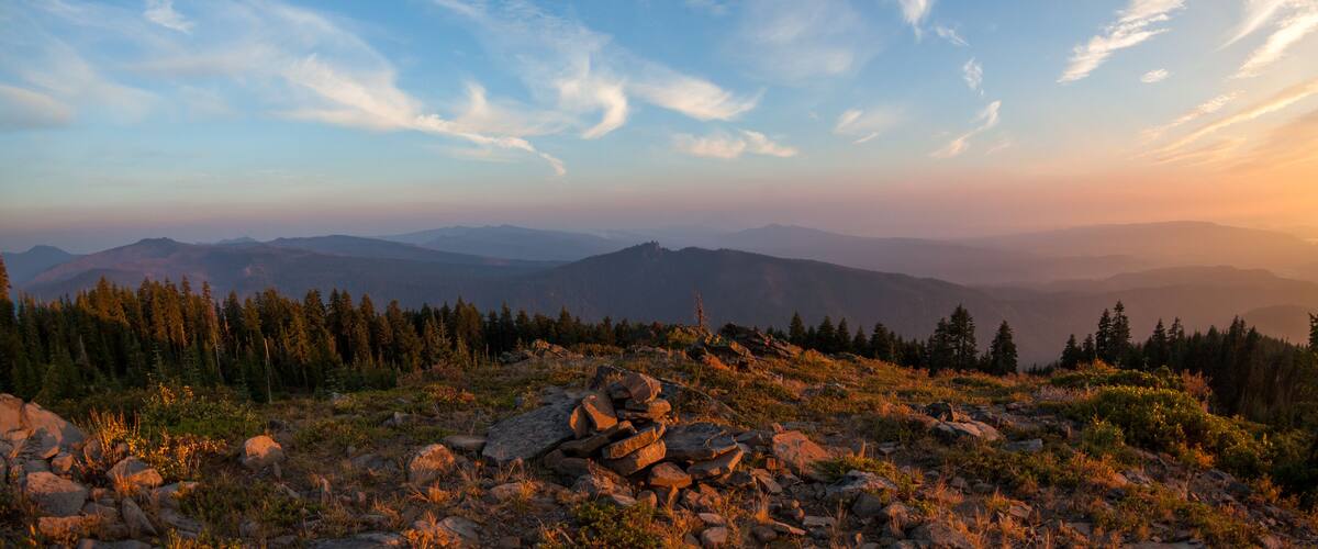 A mountainous landscape with rocky terrain, dense forests, and a clear sky with scattered clouds.