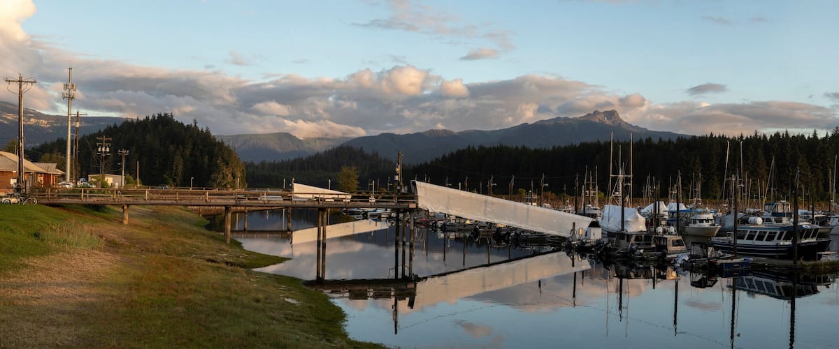 A marina with boats docked, a bridge, and mountains in the background.