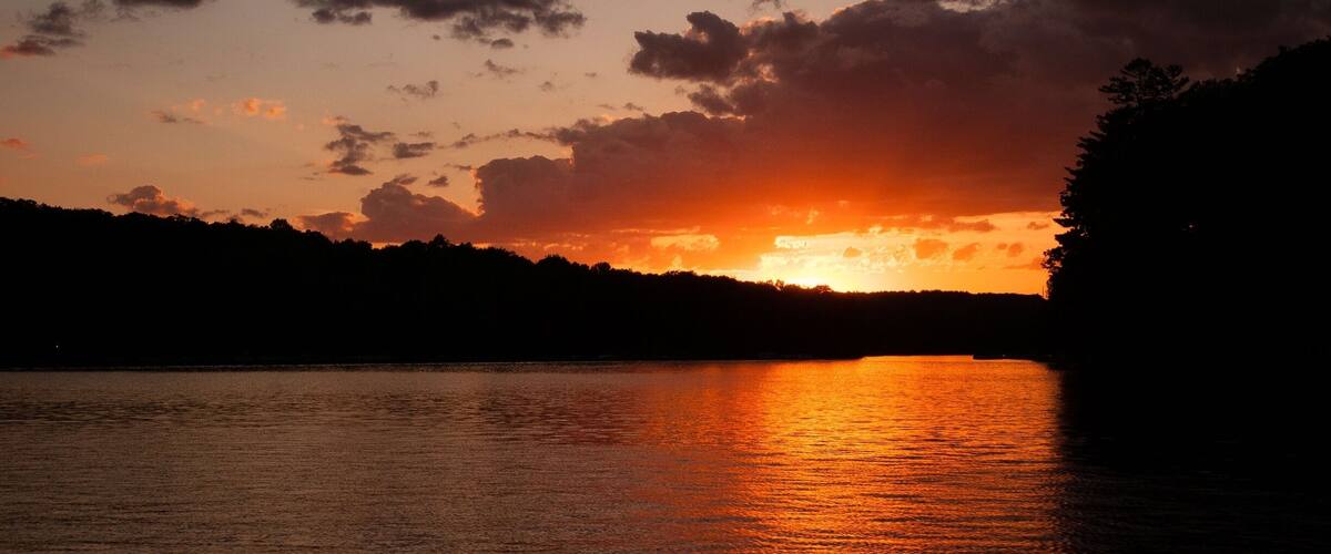 A sunset over a body of water with silhouettes of trees and a cloudy sky.