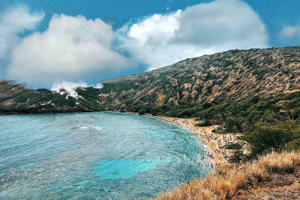 A coastal landscape with a beach, clear turquoise waters, and a mountainous backdrop.