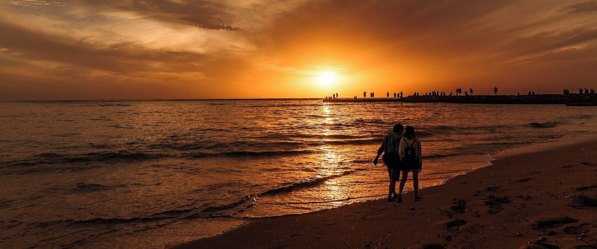 A sunset over the ocean with a beach and people walking along the shore.