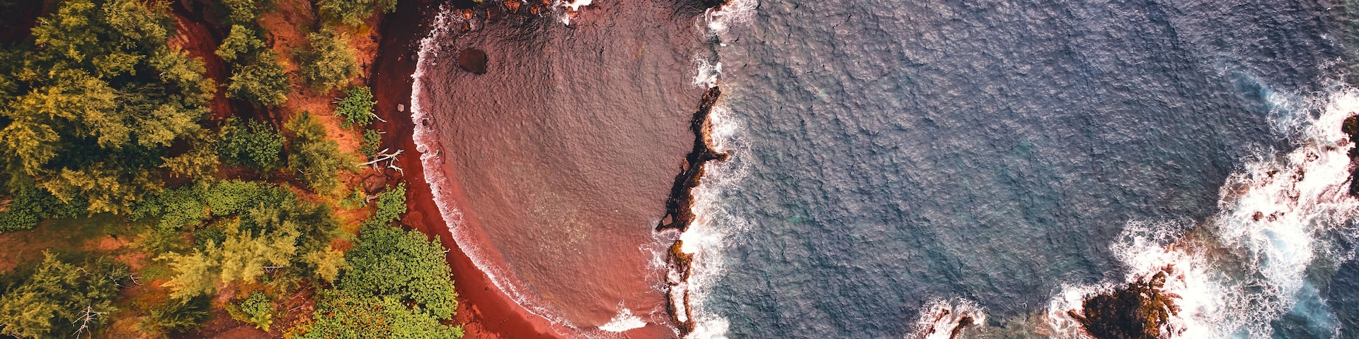 Vista aérea de una zona costera con una playa de arena roja, vegetación verde y un litoral rocoso.