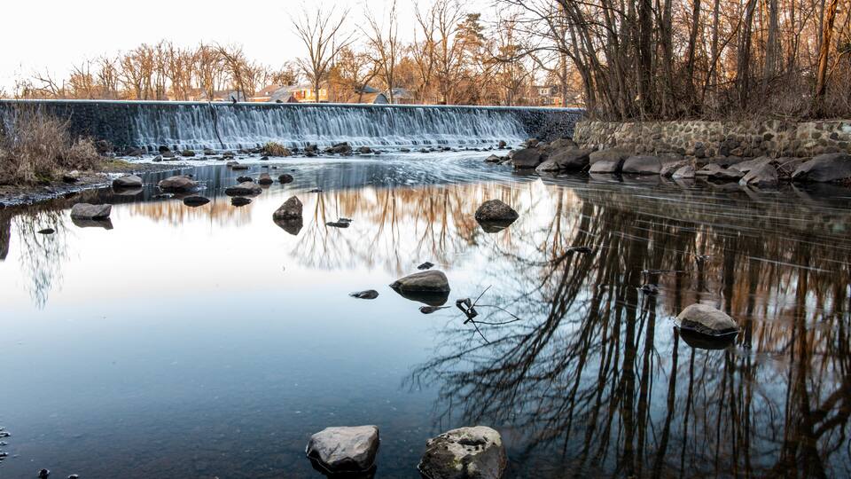 A calm river with a waterfall, surrounded by bare trees and a stone wall.