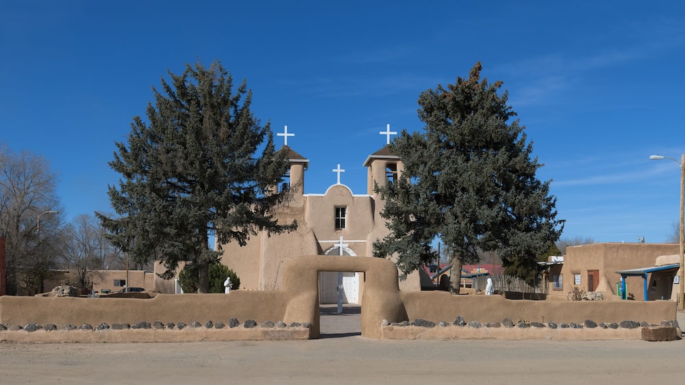 A church with a bell tower, two crosses, and a large tree in front.