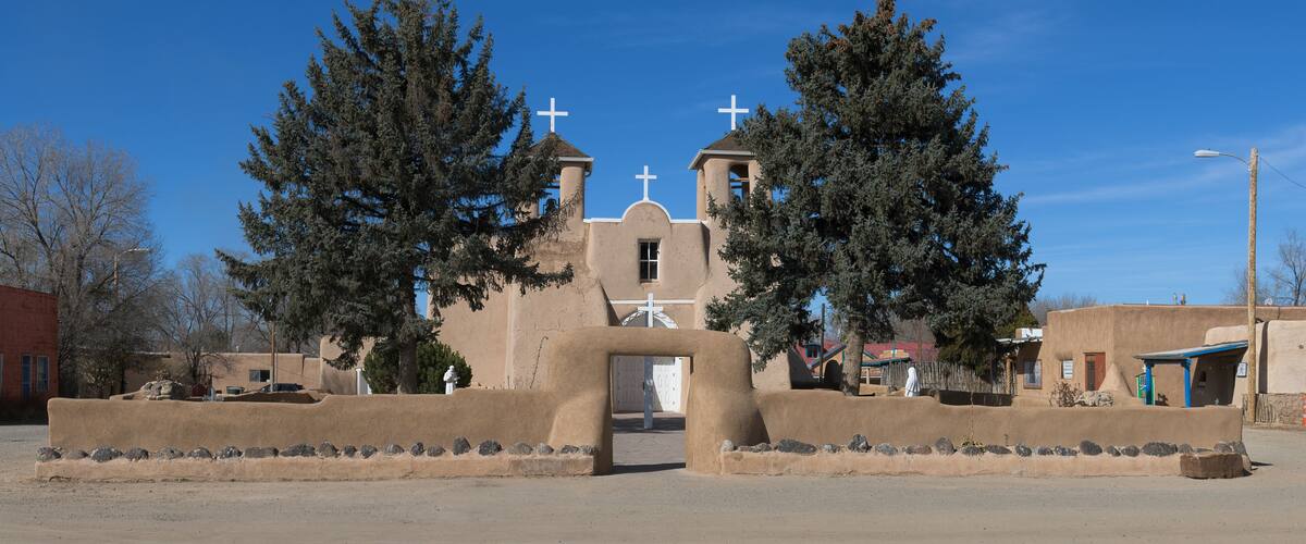 A church with a bell tower, two crosses, and a large tree in front.