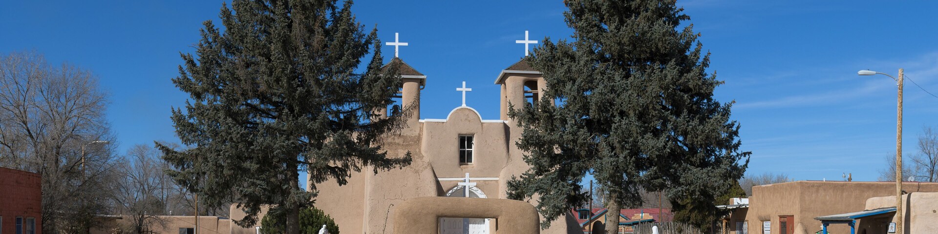 A church with a bell tower, two crosses, and a large tree in front.