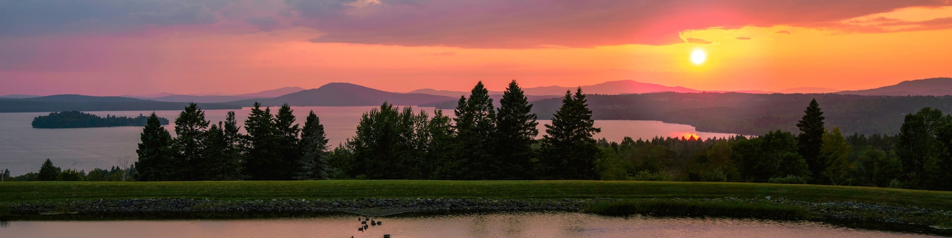 A sunset over a lake with a grassy foreground and a stone pathway.