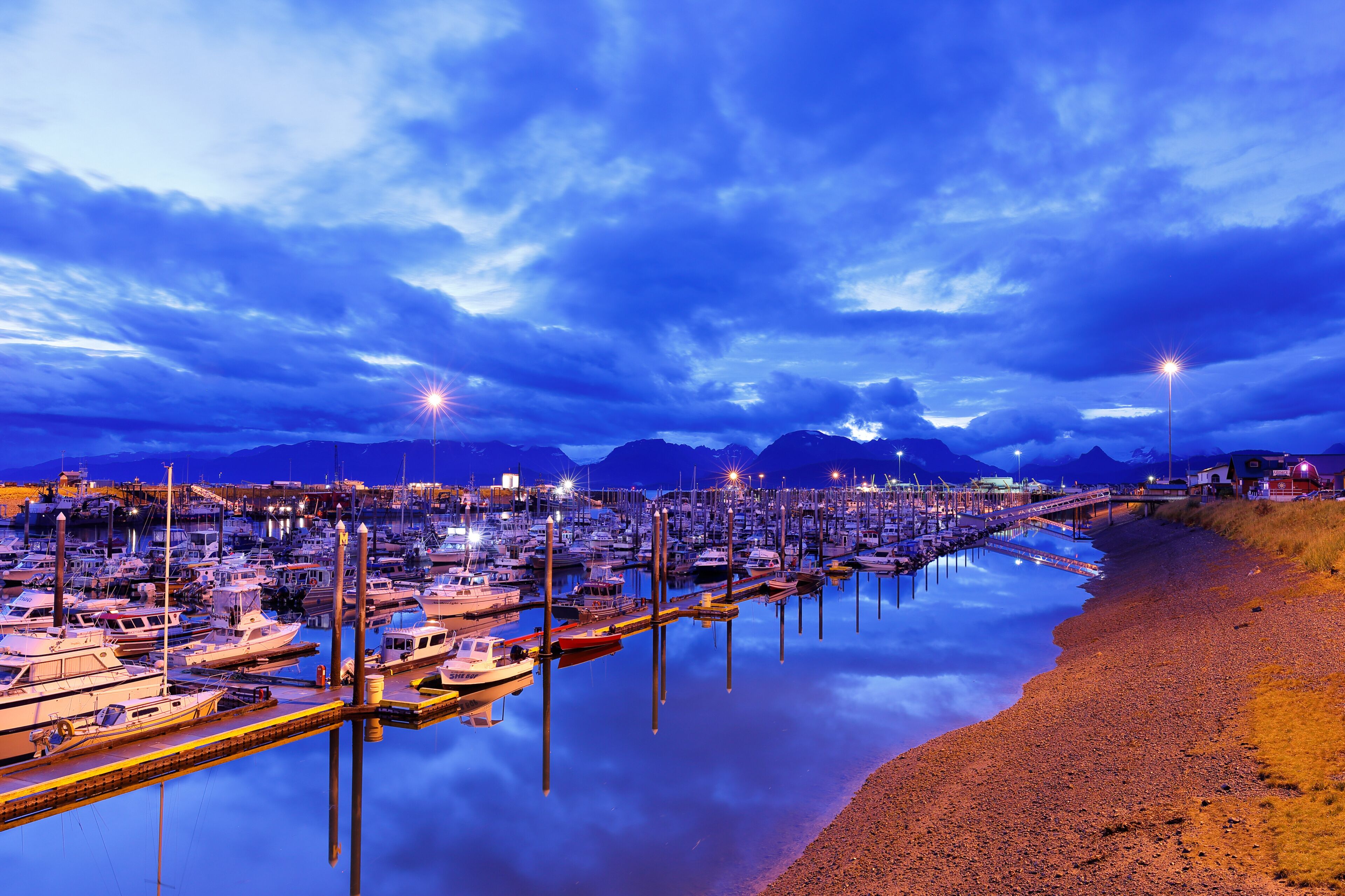 A marina with boats docked, a calm river, and mountains in the background.