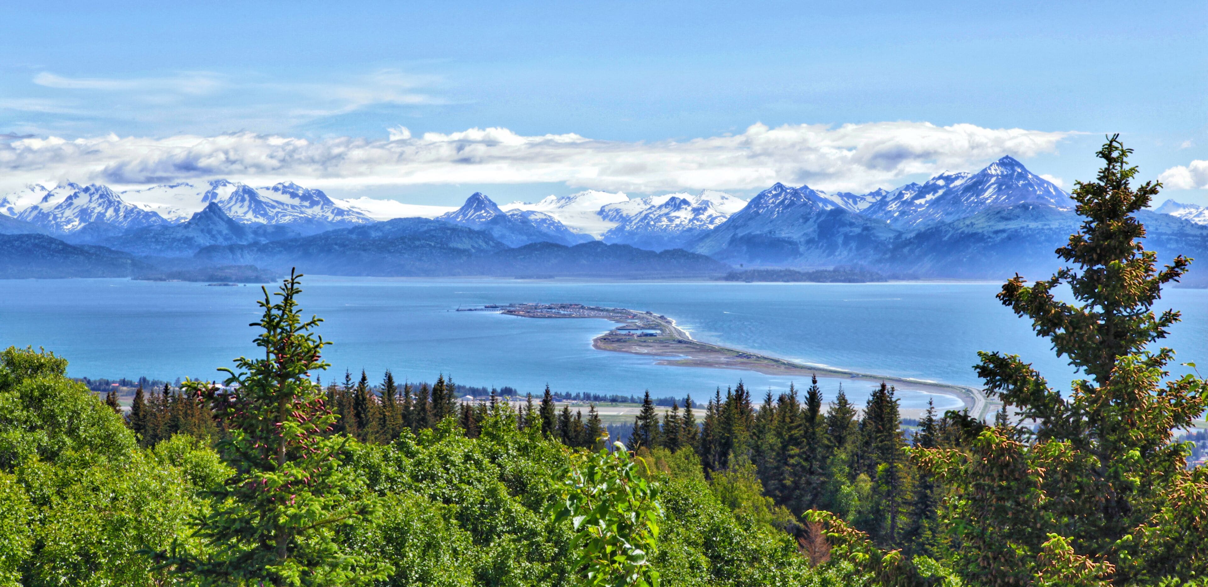 A coastal landscape with snow-capped mountains, a clear blue lake, and dense green forest.