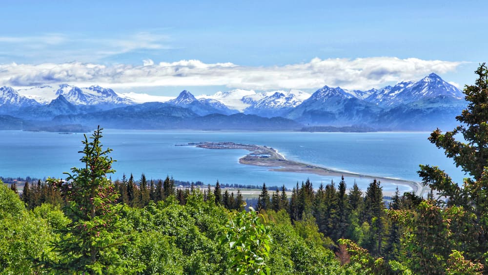 A coastal landscape with snow-capped mountains, a clear blue lake, and dense green forest.