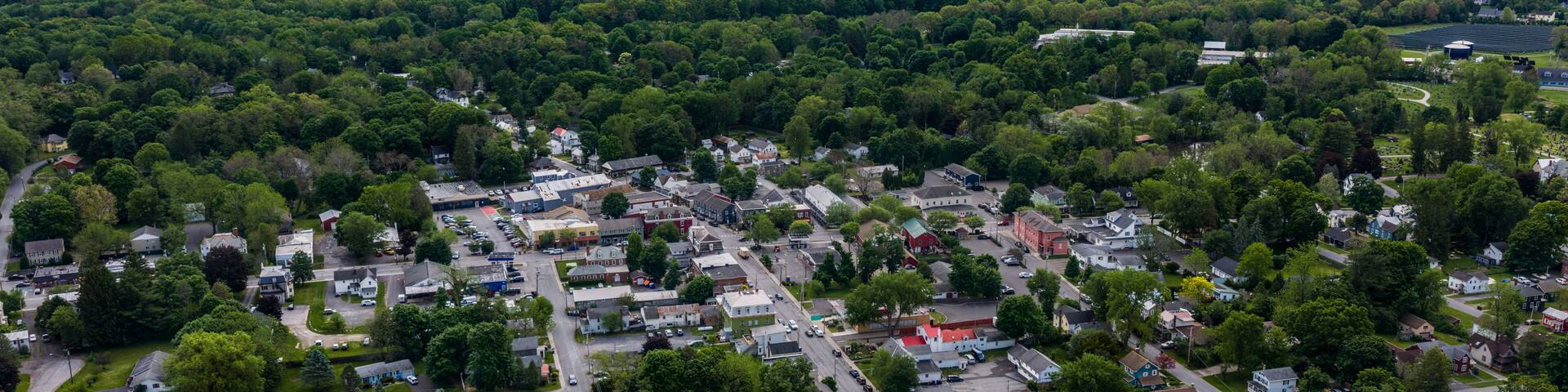 Red Hook, NY, USA - 05-20-2026 Aerial view of a small town nestled within a vast expanse of lush green forest under a partly cloudy sky.