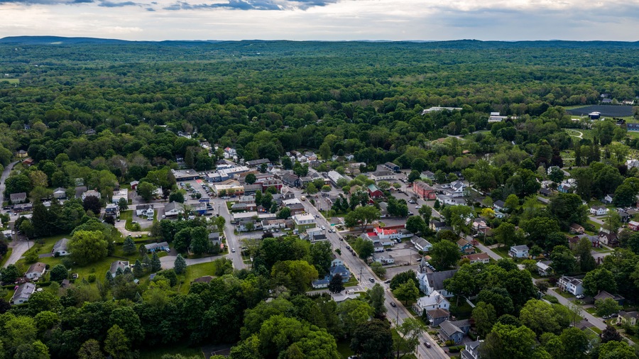 Red Hook, NY, USA - 05-20-2026 Aerial view of a small town nestled within a vast expanse of lush green forest under a partly cloudy sky.