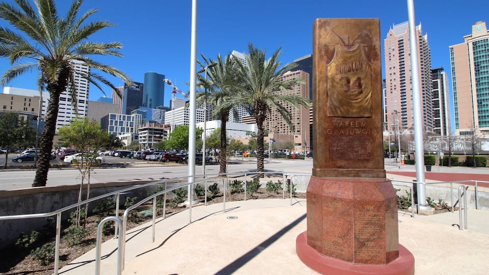 Eine bronzene Statue eines Basketballspielers mit der Trikotnummer 34, umgeben von Palmen und modernen Gebäuden im Hintergrund.