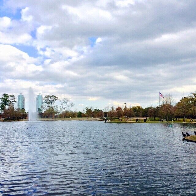 A lake with a fountain, trees, and distant buildings.