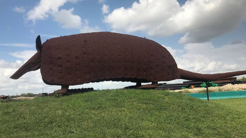 A large, textured sculpture resembling a prehistoric creature on a grassy hill with a clear sky and distant landscape.
